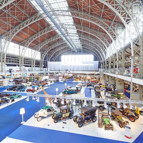 Rymligt inomhusmuseum med veteranbilar på displayen. Stort välvt tak med metallram och många utställningar med blå mattor.