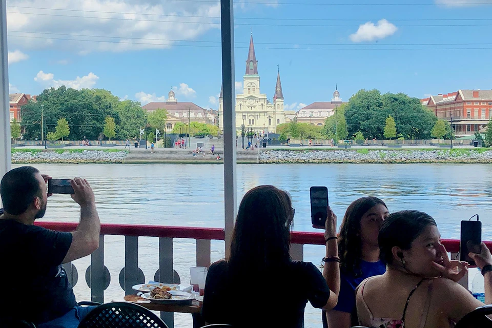 People seated on a boat, with a view of a riverside park and St. Louis Cathedral in New Orleans in the background.