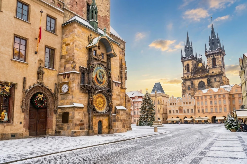 A snowy square featuring a historic clock tower and a Christmas tree, with a church in the background under a colorful sky.