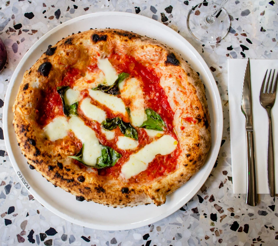 Neapolitan-style pizza with mozzarella, tomato sauce, and basil on a white plate, next to a fork and knife on a terrazzo table.