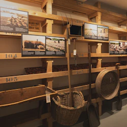 En museiutställning med trähyllor med verktyg, korgar och informationspaneler om historiska jordbruksmetoder.