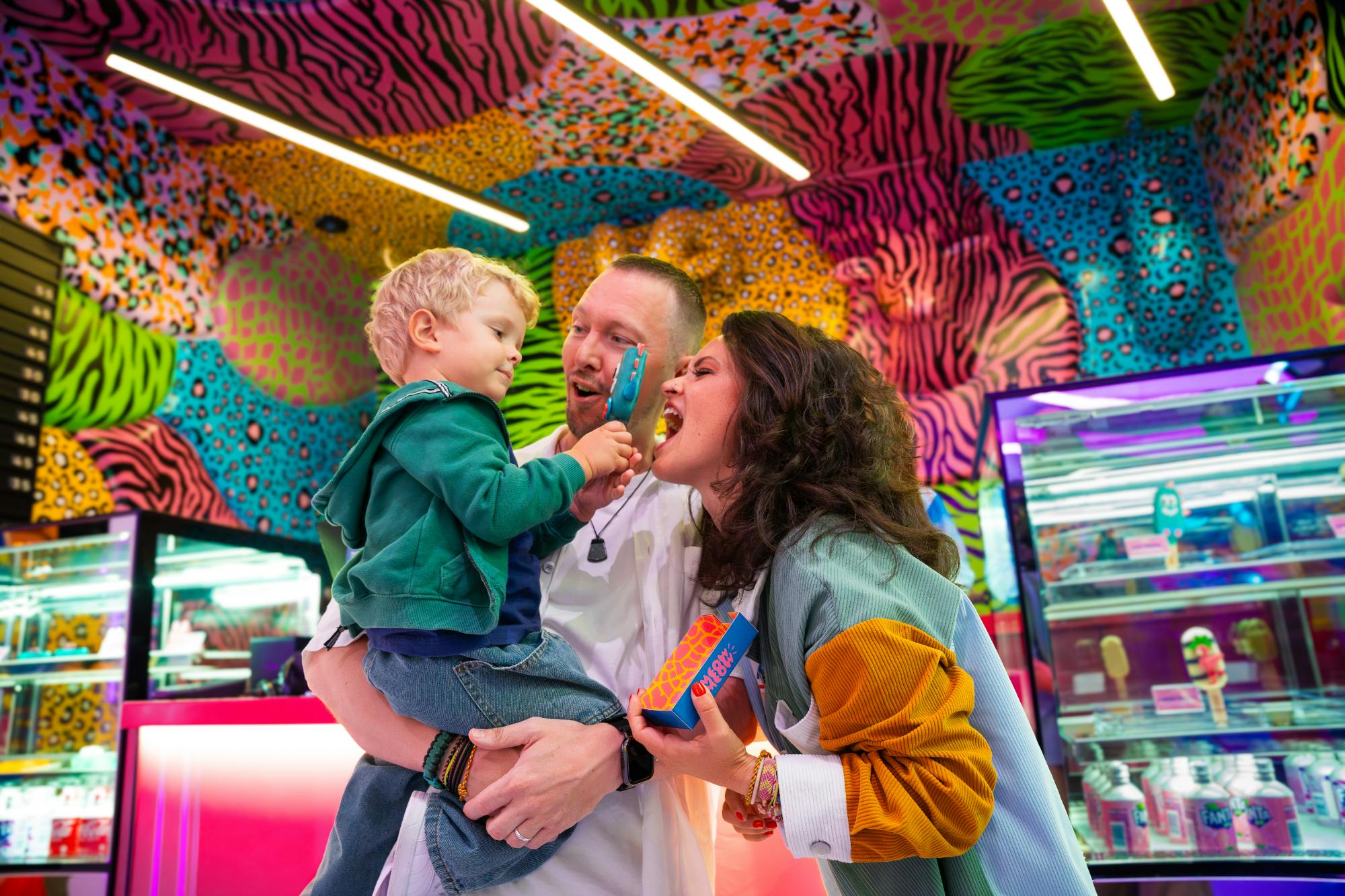 Rodzina w kolorowym pokoju; ojciec trzymający malucha, który podaje kobiecie loda, podczas gdy ona żartobliwie udaje, że go je.