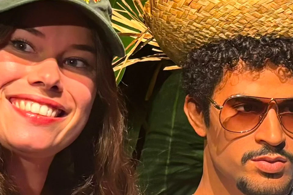 Two people in front of a lush green background, one taking a selfie and the other wearing sunglasses and a straw hat.