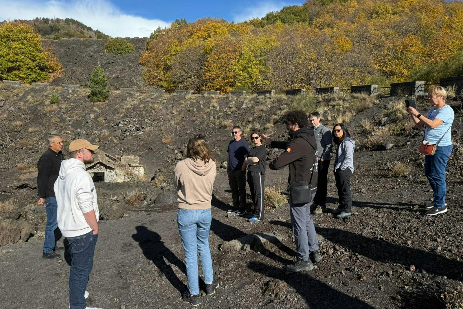 Un gruppo di persone in piedi all’aperto su un terreno roccioso, vicino ad alberi con foglie autunnali e una piccola struttura sullo sfondo.