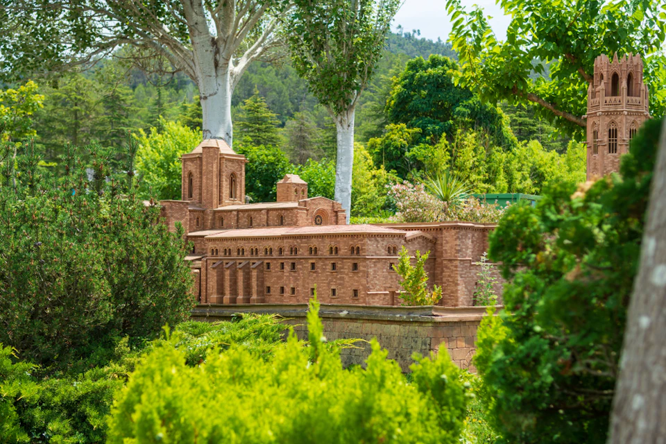 A scale model of a historic brick building is surrounded by lush greenery and trees with a forested hillside in the background.
