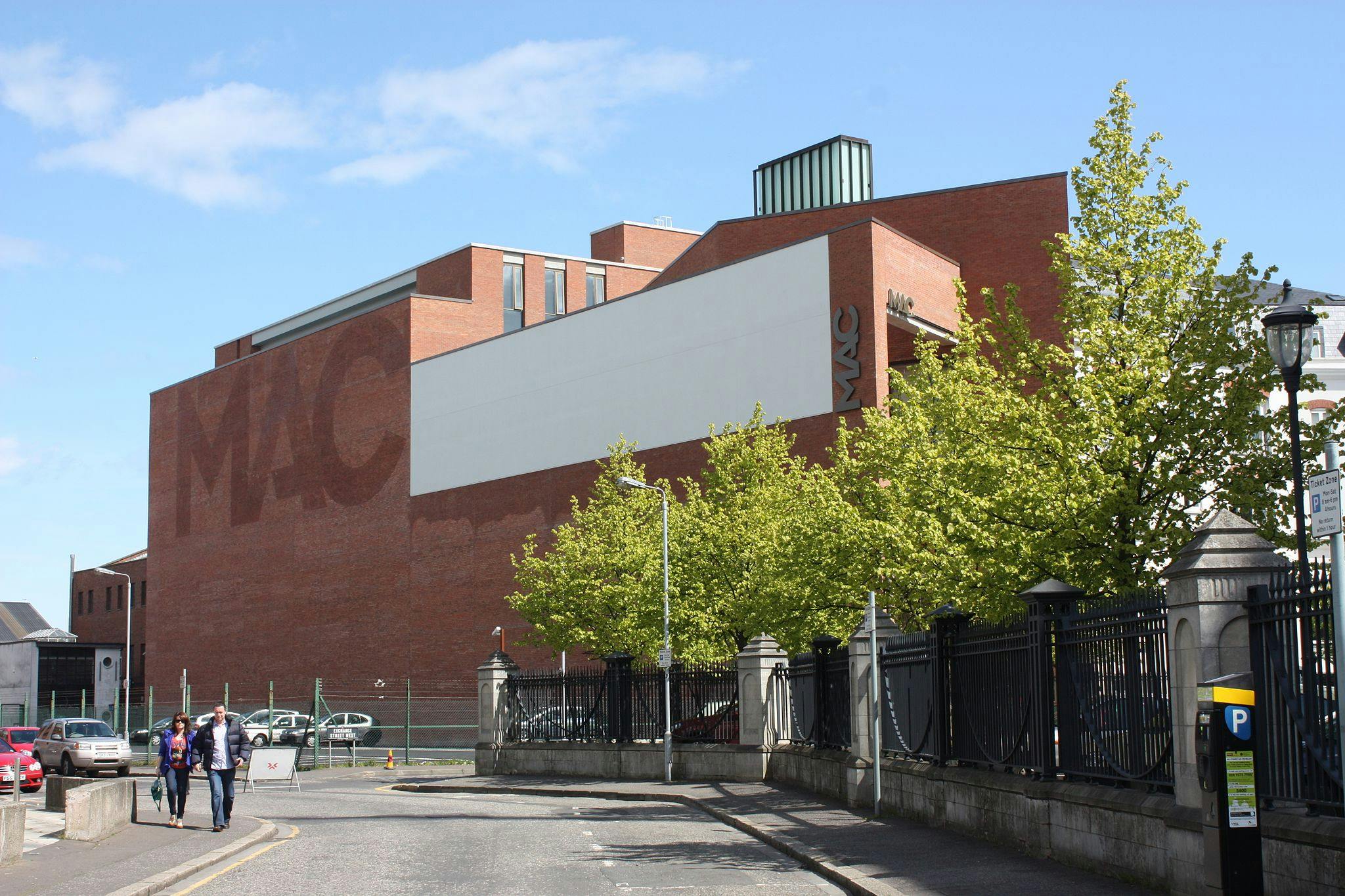 Two people walking on a sidewalk near a large brick building labeled "MAC" with trees and parked cars on a sunny day.