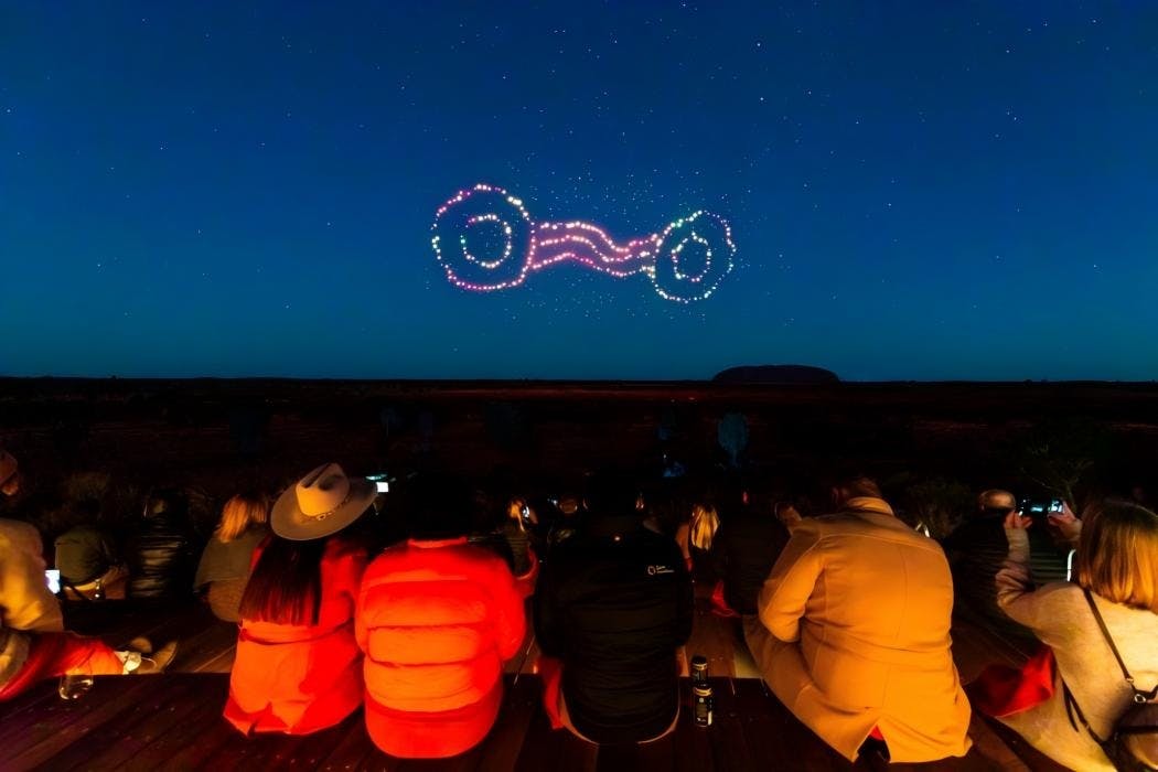 A group of people watch a drone light show forming interconnected rings in the night sky.