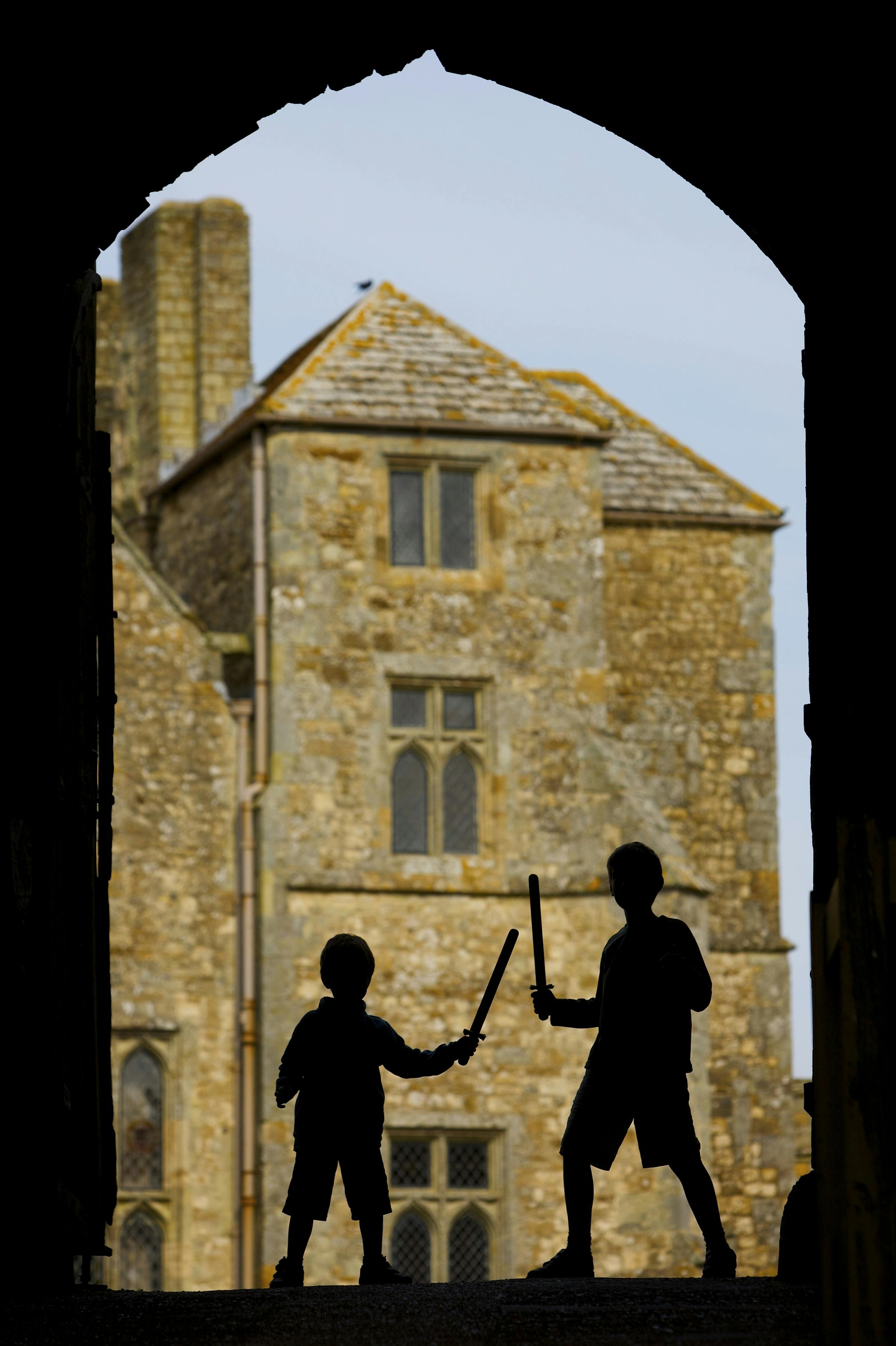 两个孩子拿着玩具剑，背景是一座古老的石头建筑，窗户是拱形的，屋顶是瓷砖。