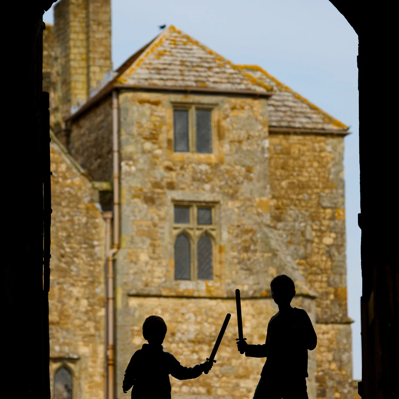 Two children with toy swords silhouetted against the backdrop of an old stone building with arched windows and a tiled roof.