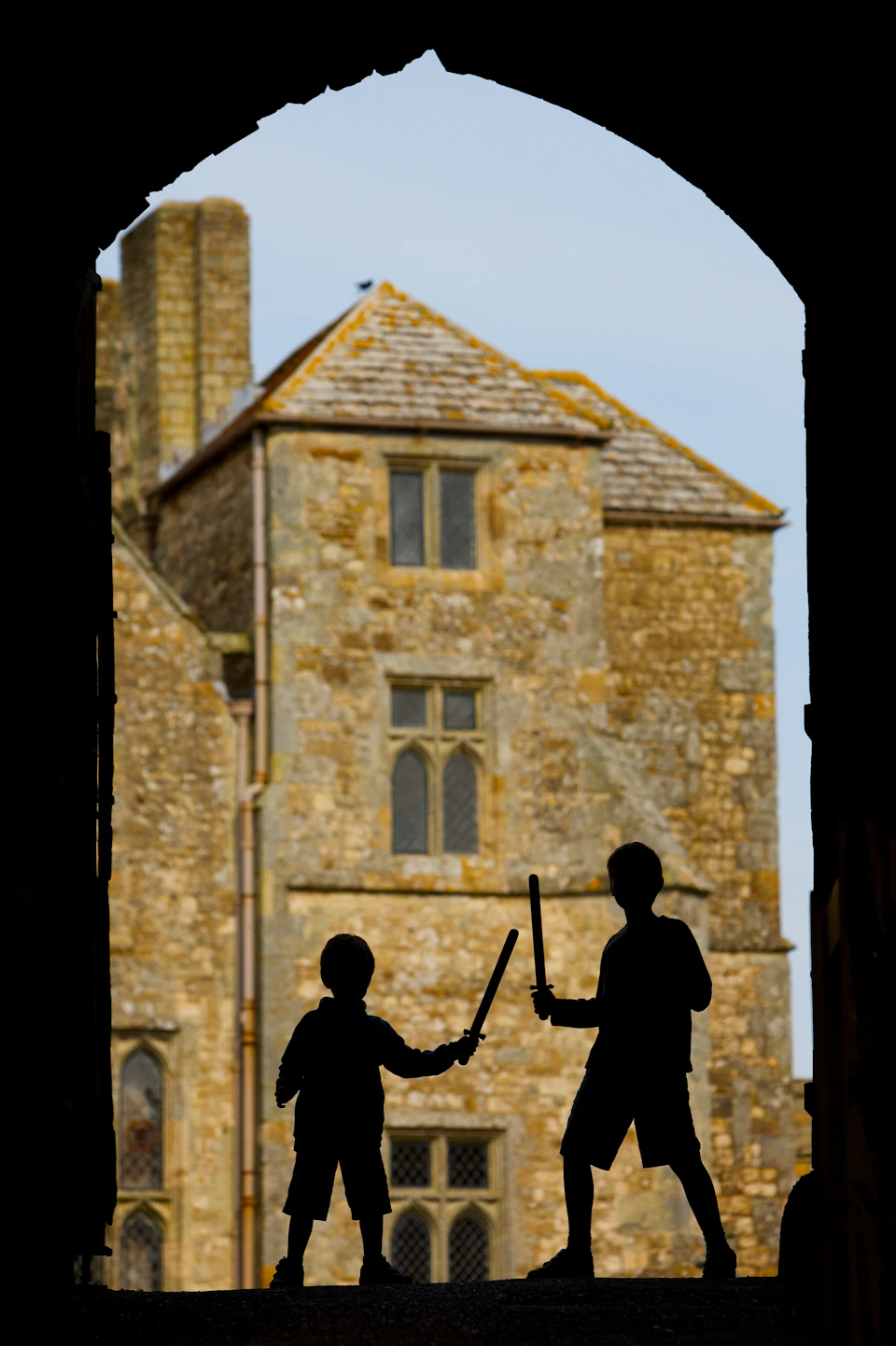 Two children with toy swords silhouetted against the backdrop of an old stone building with arched windows and a tiled roof.