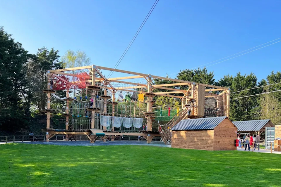 Outdoor adventure course with wooden platforms, ropes, nets, zip lines, and a small wooden shed; surrounded by grass and trees.