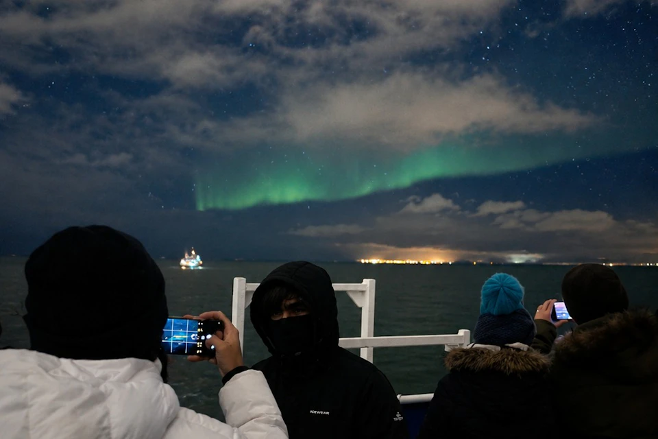 People on a boat photographing the northern lights under a cloudy, starry night sky with distant lights on the horizon.