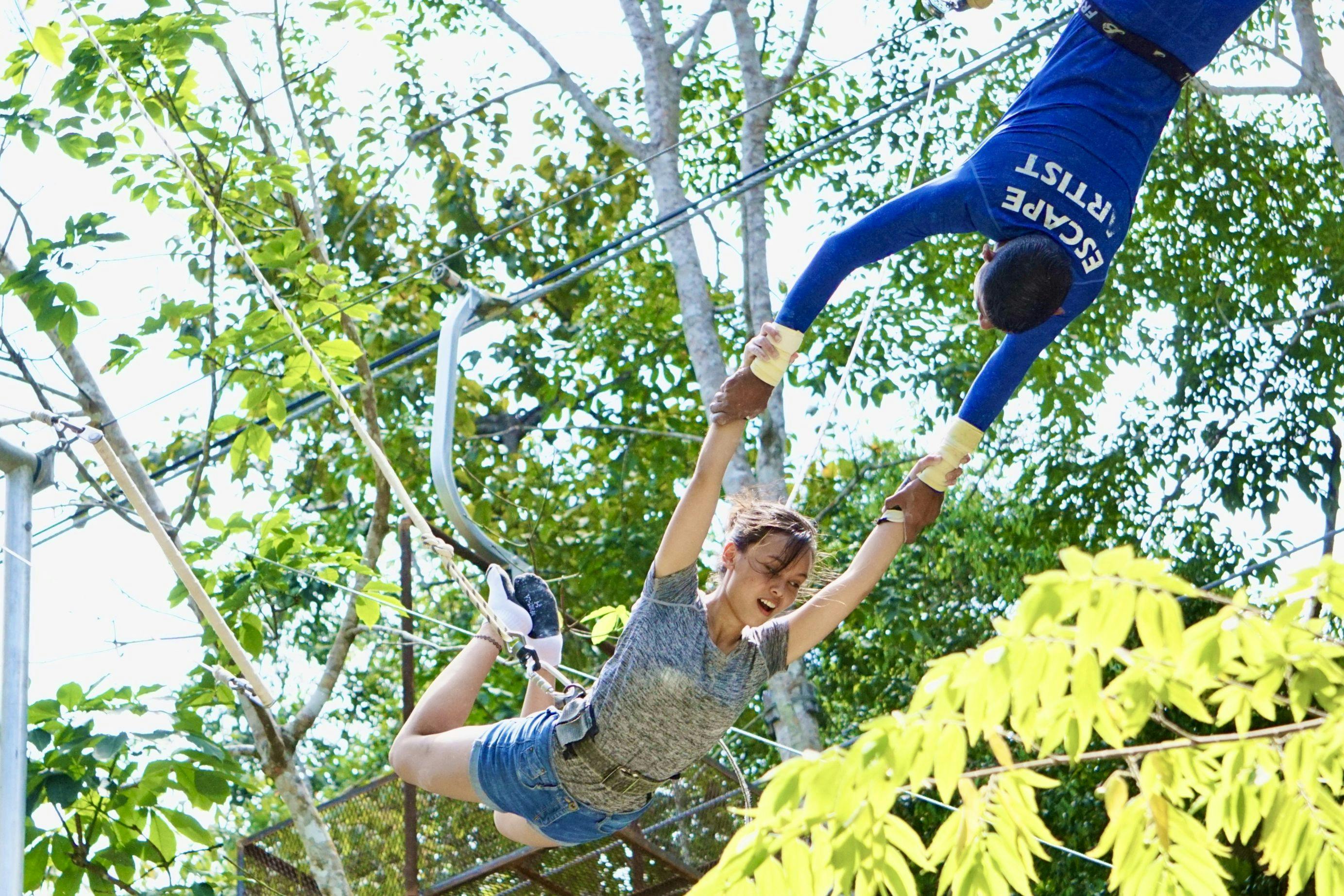 A woman swinging on a harness while a person in a blue shirt assists her in a lush outdoor setting.