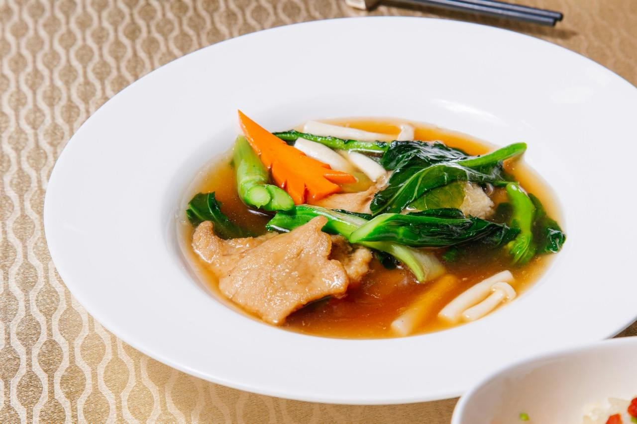 A white bowl containing a soup with vegetables, meat slices, and noodles in a brown broth, served on a patterned tablecloth.