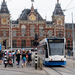 En spårvagn märkt "26 Ijburg" framför Amsterdams centralstation med människor som går och en röd tegelbyggnad i bakgrunden.