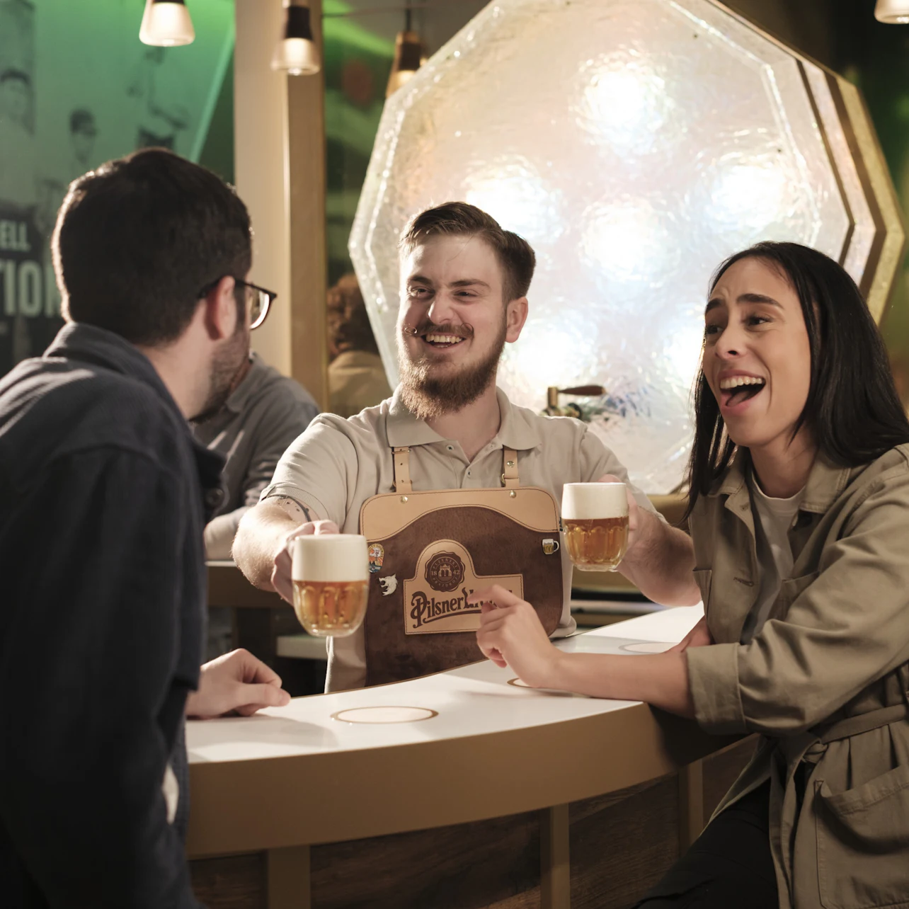 Bartender serves beer to two laughing patrons at a well-lit bar with a large textured light fixture in the background.