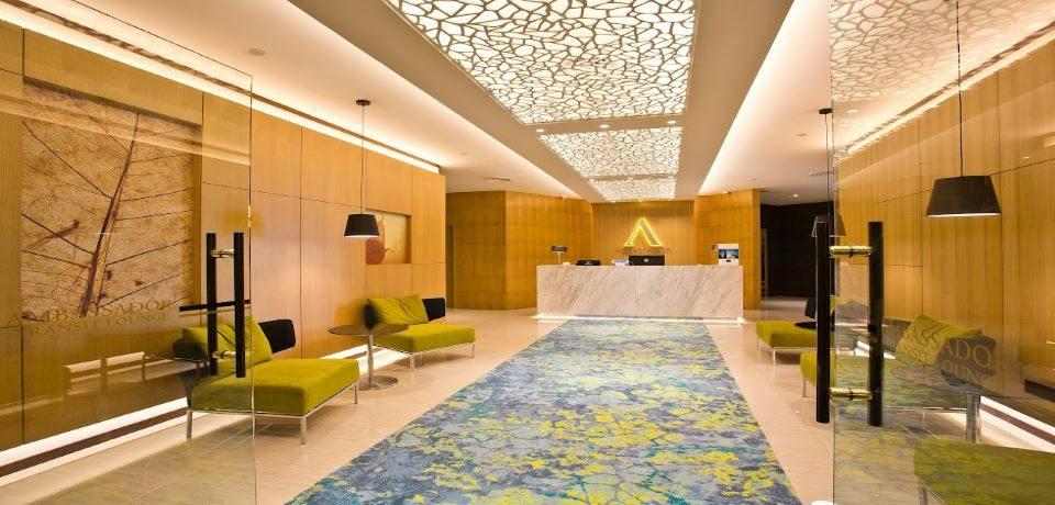 A modern hotel lobby with a marble reception desk, wooden walls, green chairs, and a patterned blue carpet under artistic lighting.