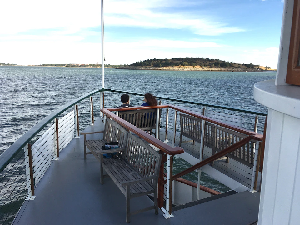 Two people sitting on a bench at the deck of a boat, with a distant island and calm waters under a partly cloudy sky.