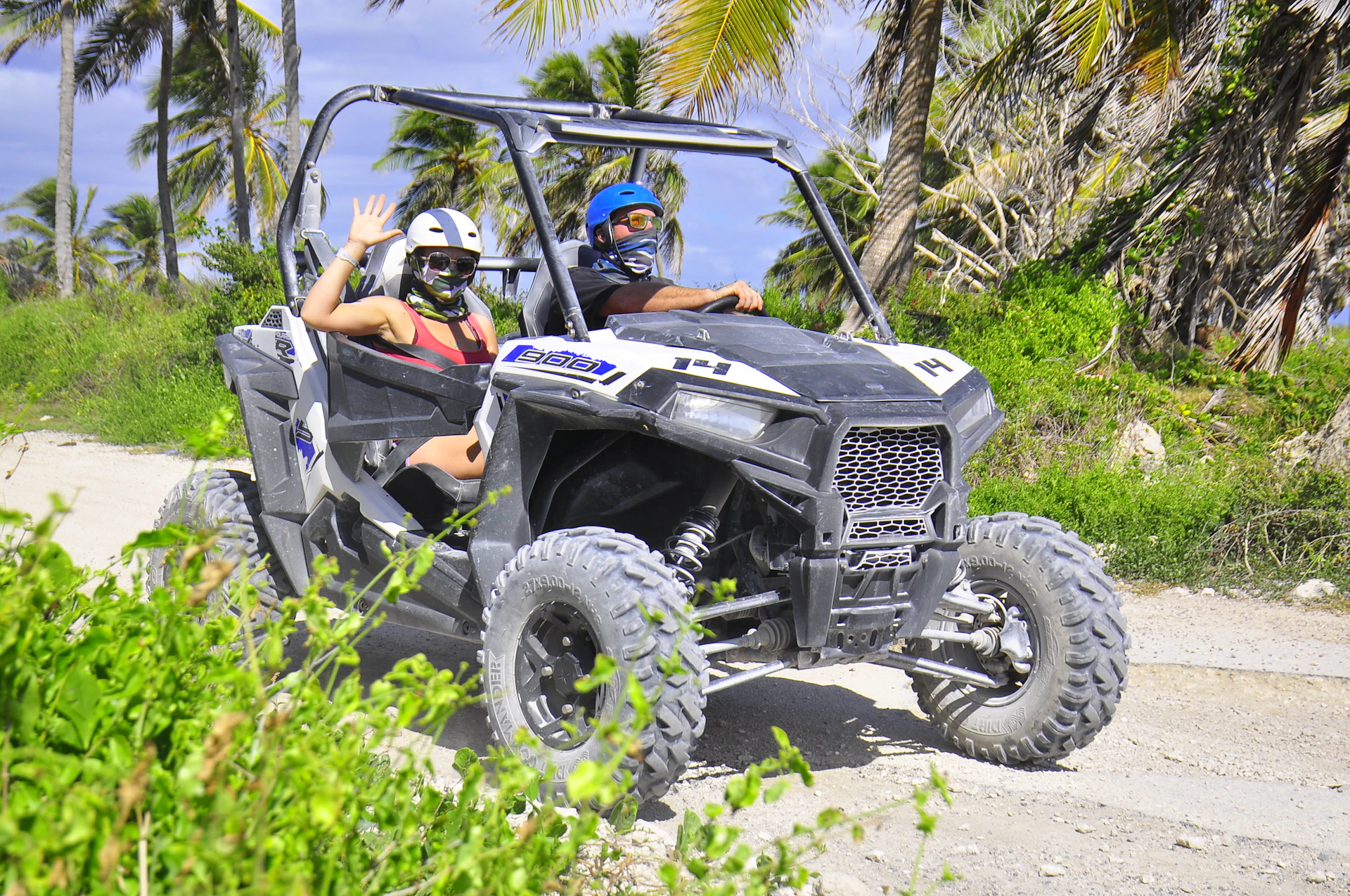 Two people wearing helmets and masks ride a dune buggy on a sandy path lined with palm trees. One person waves.