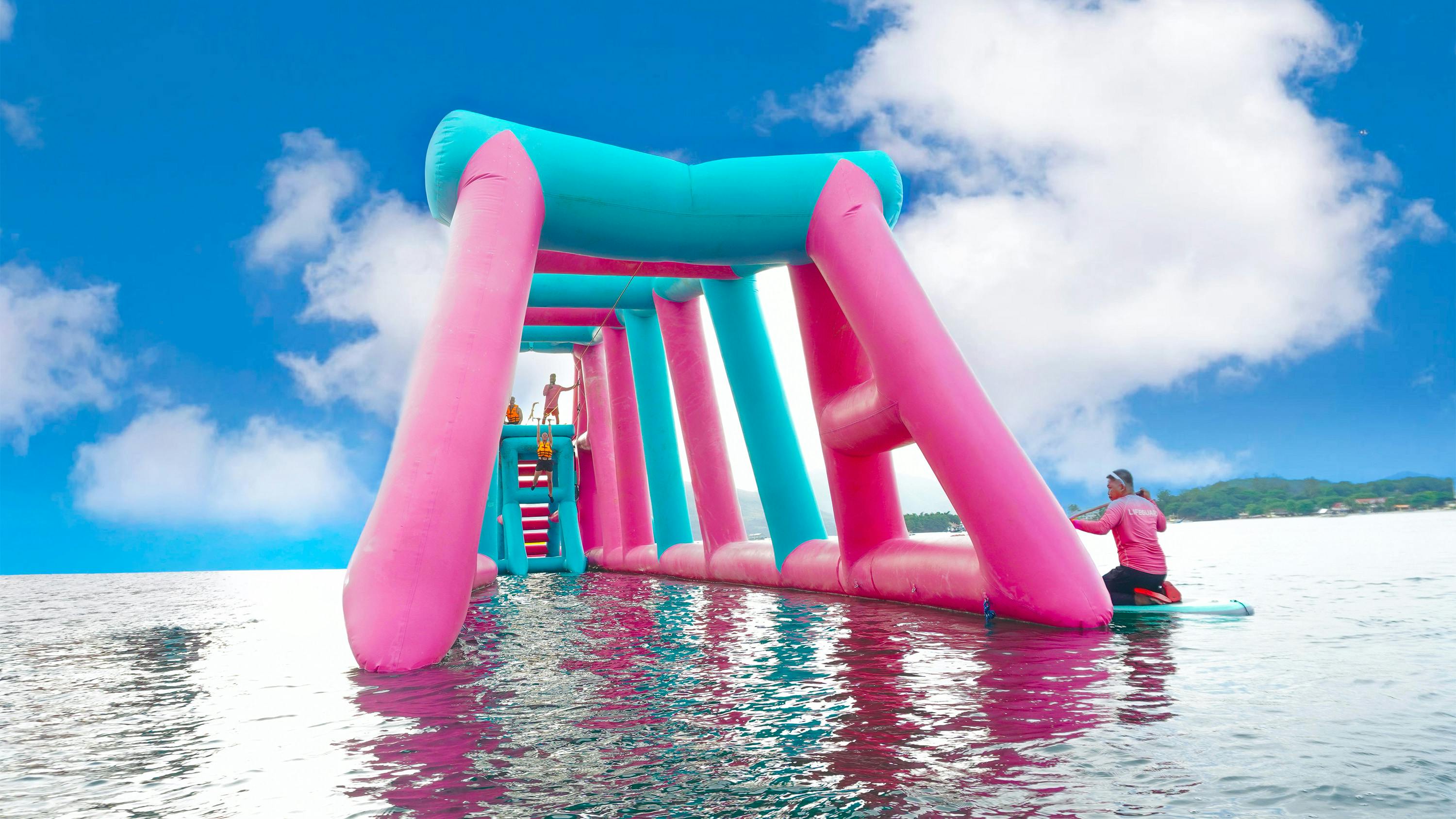 Large pink and blue inflatable structure on water under a bright sky, with people climbing and standing on it.