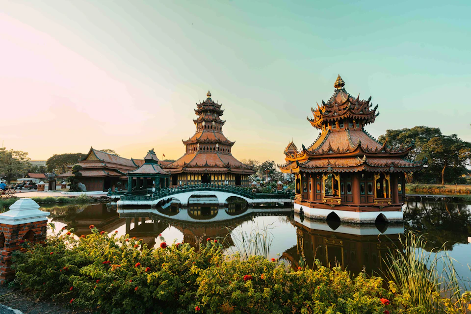 Ornate pagodas with tiered roofs beside a tranquil pond, surrounded by lush greenery and flowers, under a clear sky.