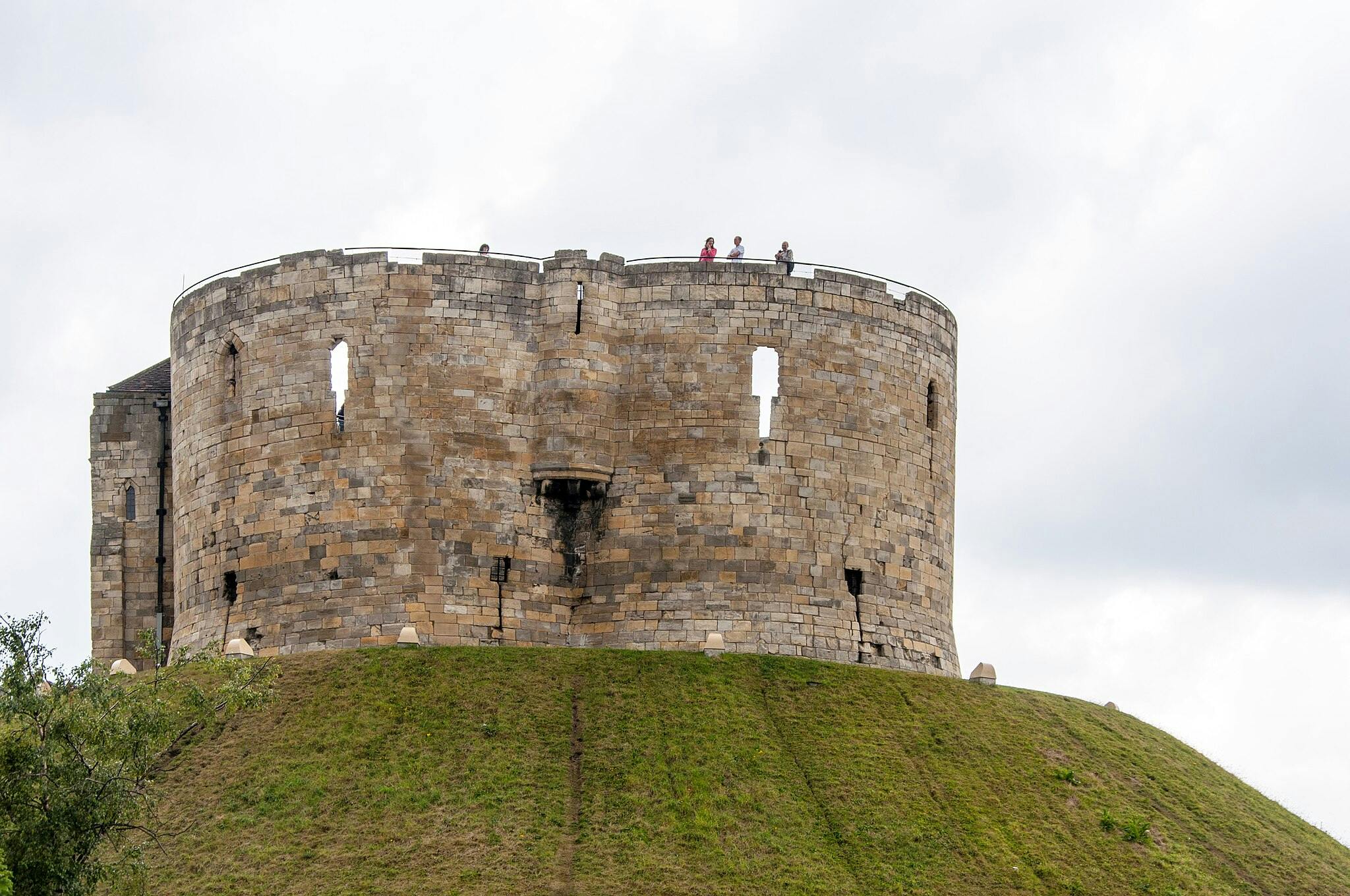 一座石头堡垒塔位于草山顶上，顶部站着四个人，在多云的天空下可见。