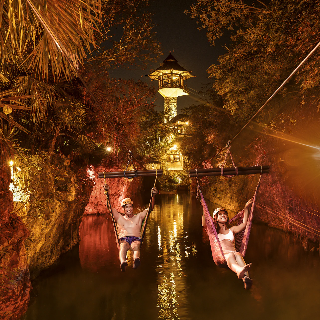 Two people zip-line over a river at night, surrounded by illuminated rocks and trees. A lit tower is visible in the background.