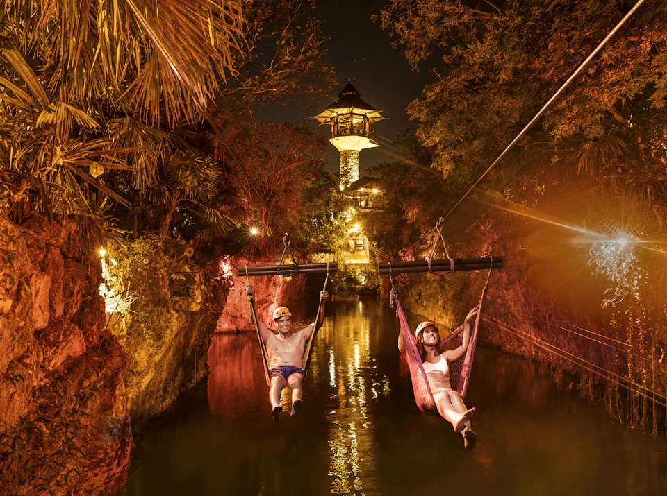 Two people zip-line over a river at night, surrounded by illuminated rocks and trees. A lit tower is visible in the background.