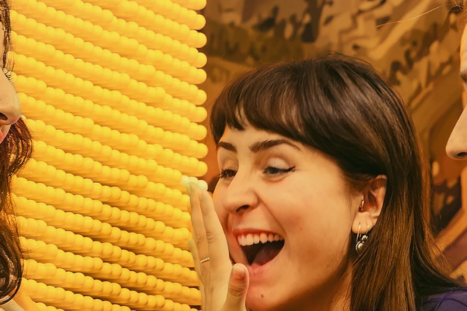 Three women excitedly greet each other indoors against a yellow textured background.