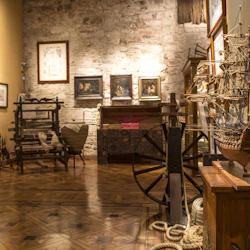 Ett museum med ett modellskepp, antika möbler, inramade bilder och en snurrande hjul på ett trägolv.