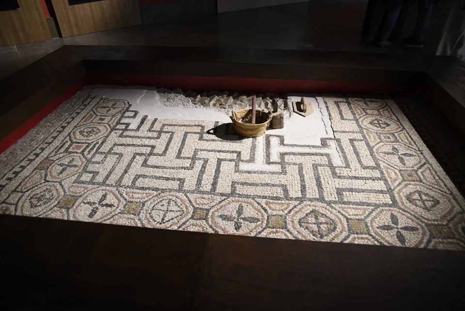 A restored ancient mosaic floor with geometric patterns, featuring a wooden bucket and tools placed on it.