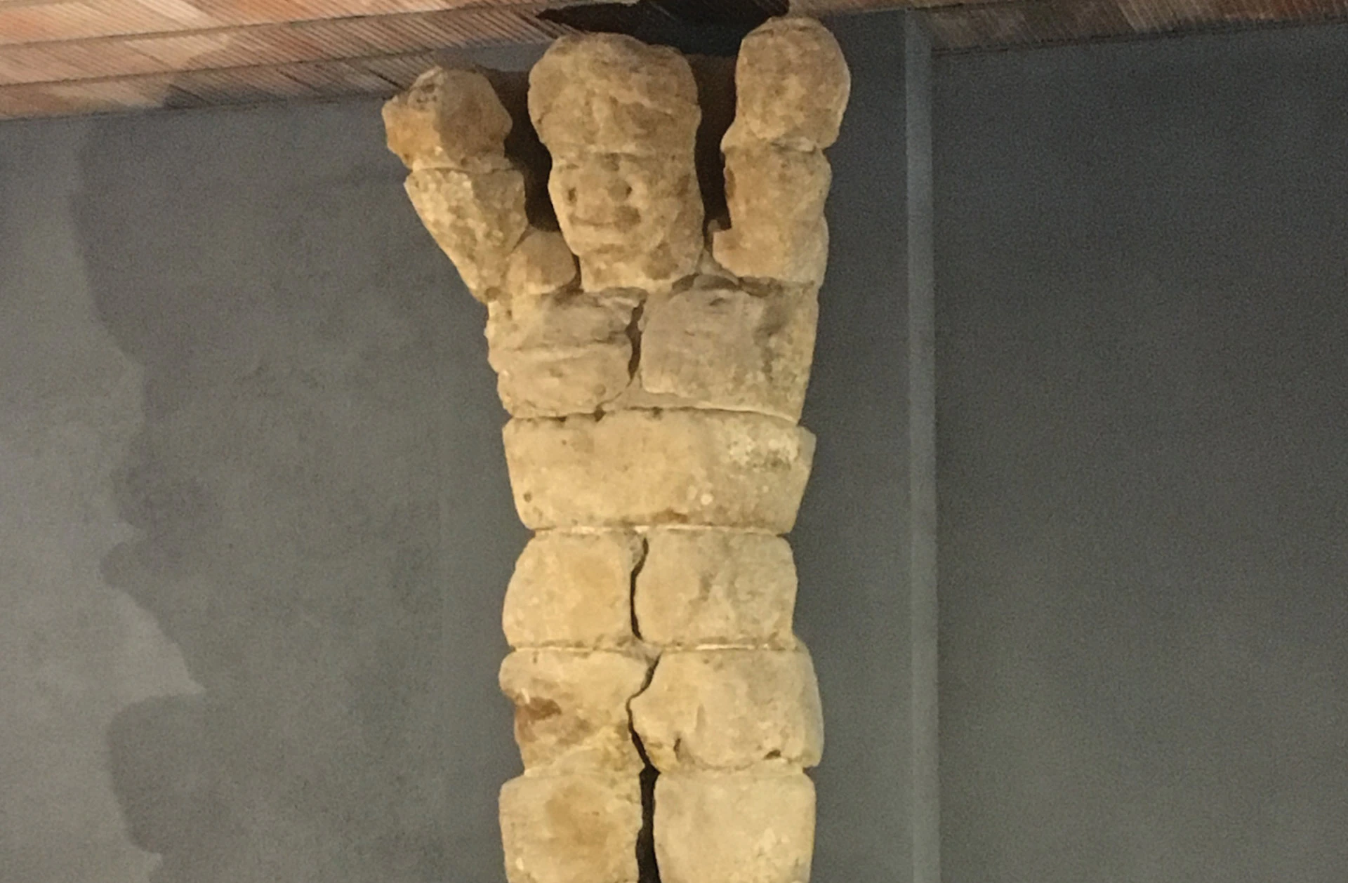 A sculpture made of stacked, irregular stone blocks resembling a human figure with raised arms against a plain wall.
