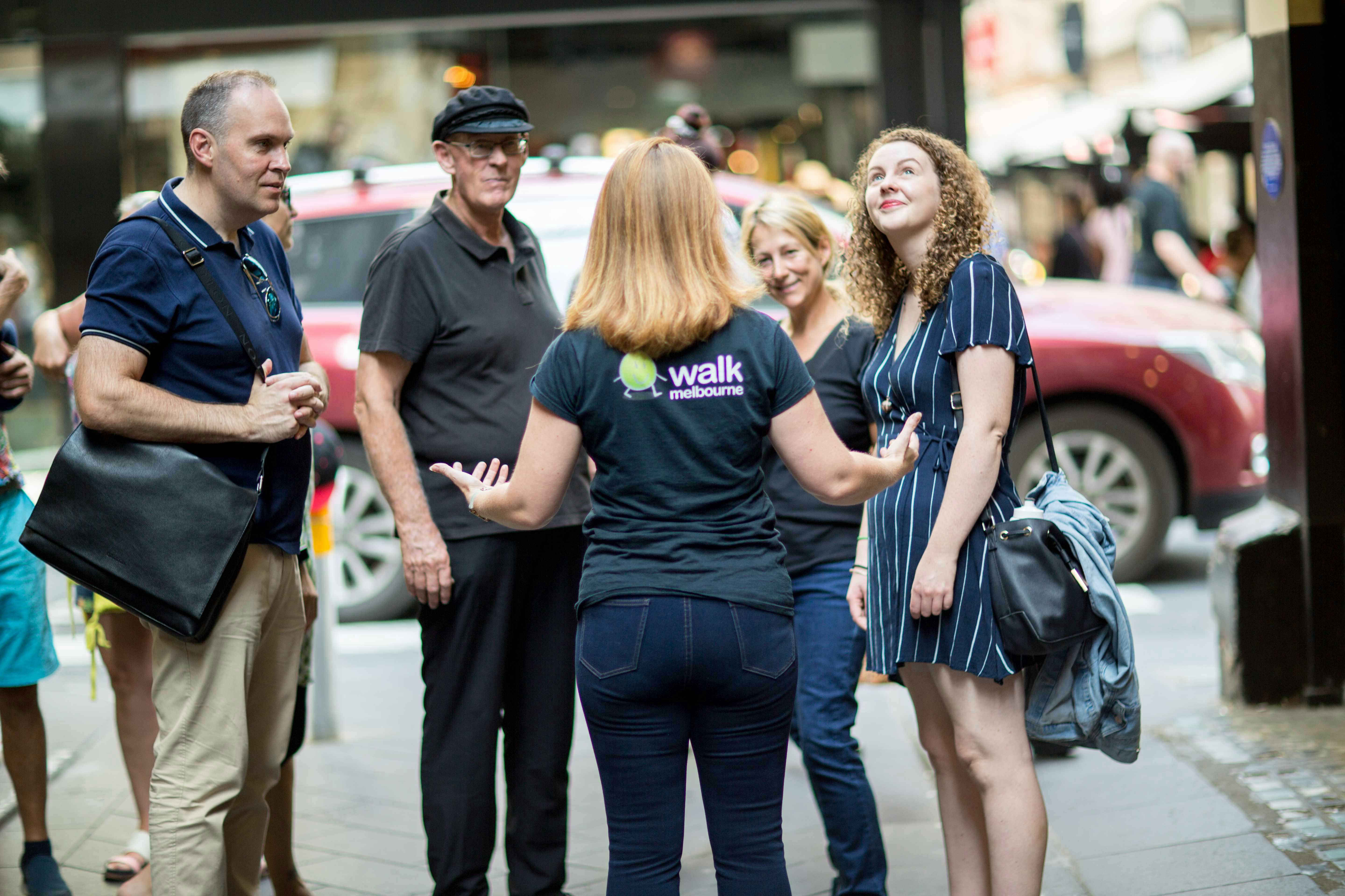 Group of people standing outdoors, engaged in conversation. One person wears a "walk melbourne" shirt. A red SUV is in the background.