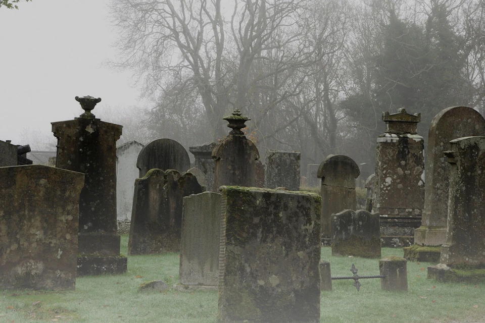 A misty graveyard with old, moss-covered gravestones and bare trees in the background.