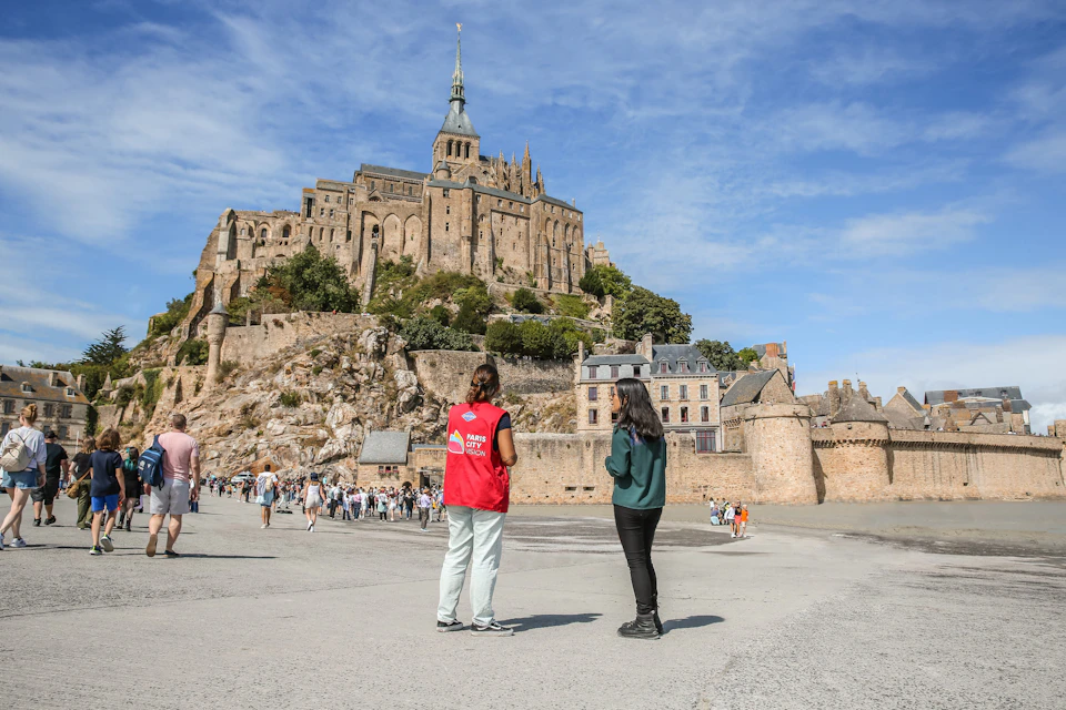 Two people stand in front of Mont Saint-Michel, a historic abbey on a hill, with crowds and stone buildings in the background.