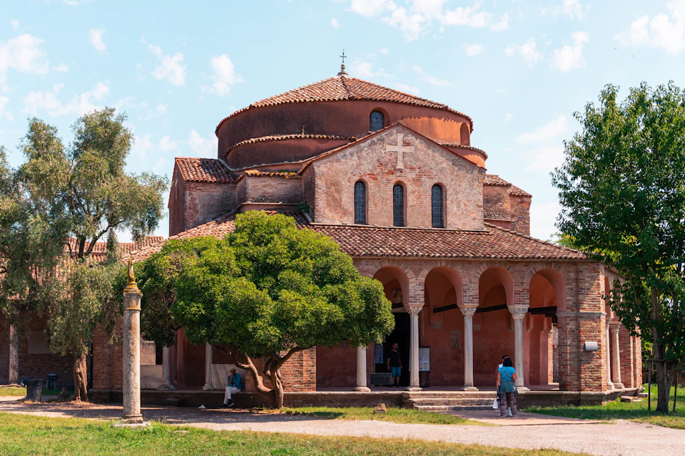 A historic brick church with a tiled roof, large arched entryway, and a cross above the entrance. People are visible near the entrance.