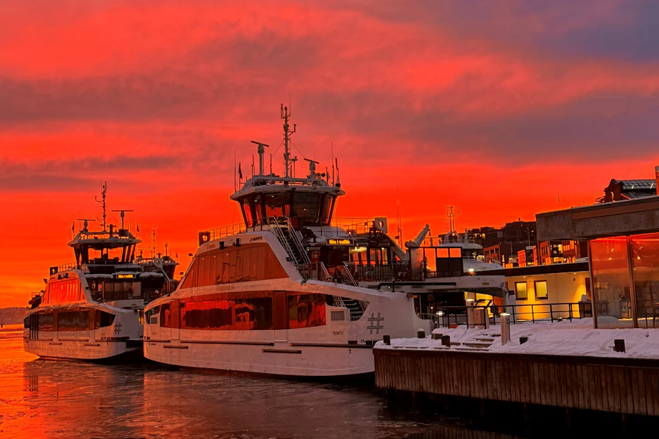 A docked ferryboat at sunset with a vibrant red and orange sky, accompanied by nearby structures and other boats.