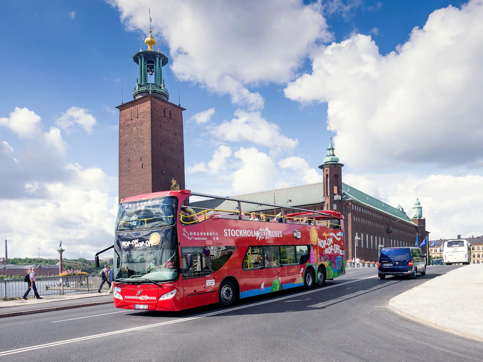 A red double-decker tour bus drives past Stockholm City Hall under a blue sky with scattered clouds.