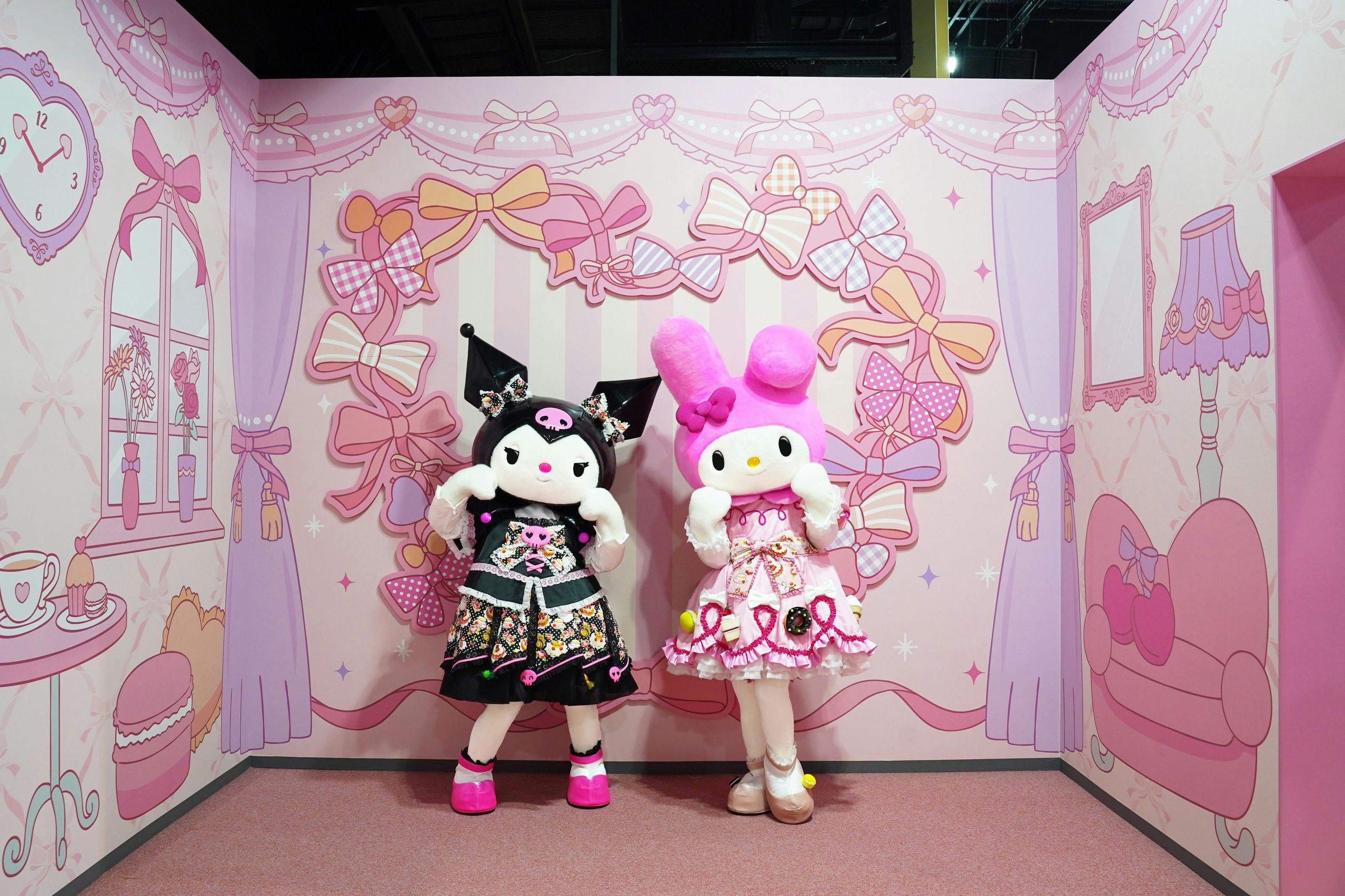 Two costumed characters in a pink, bow-themed setting, one in black attire with ears and the other in pink with a bow.