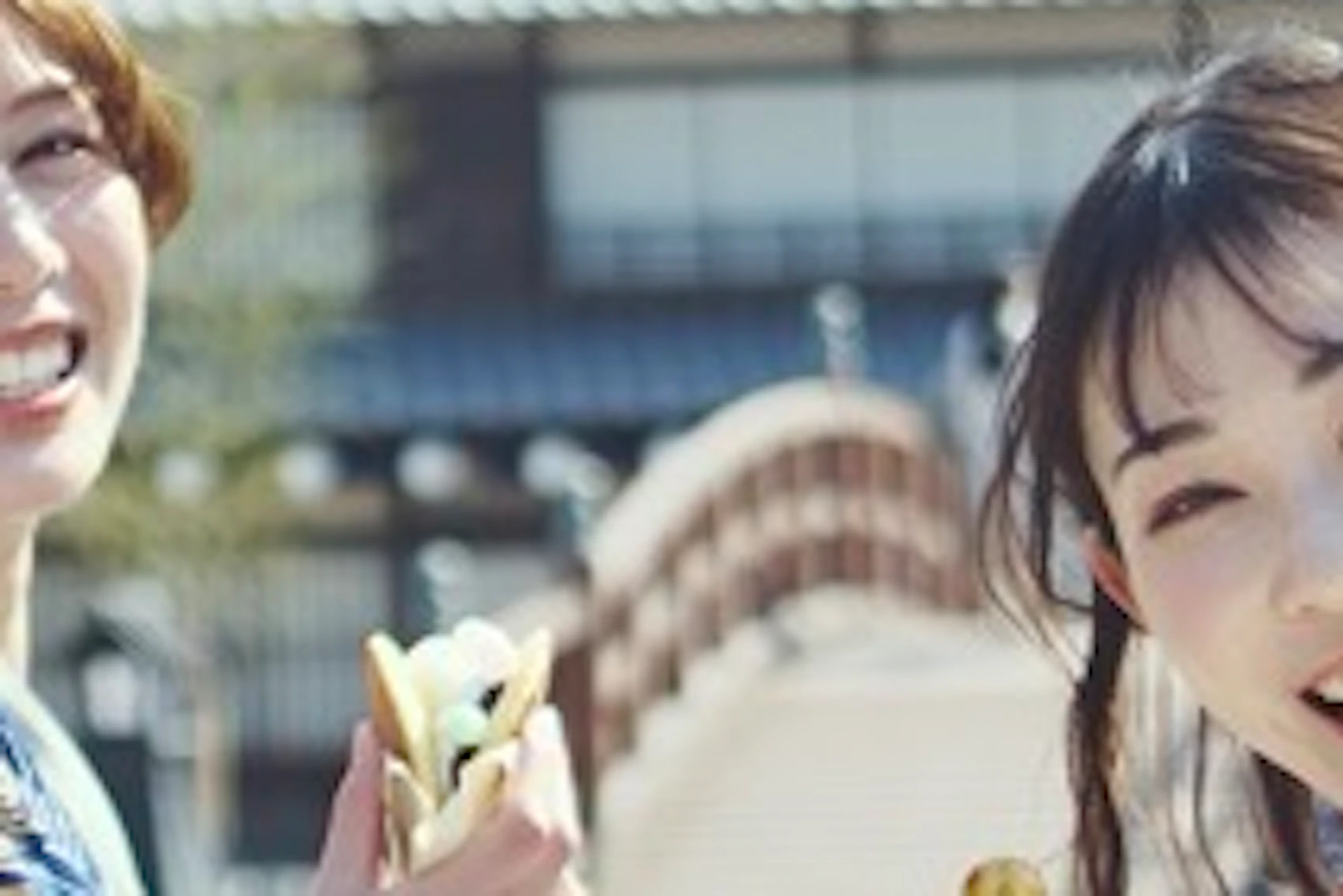 Two women in traditional Japanese attire smile and hold skewers of food, standing on a street with wooden buildings.