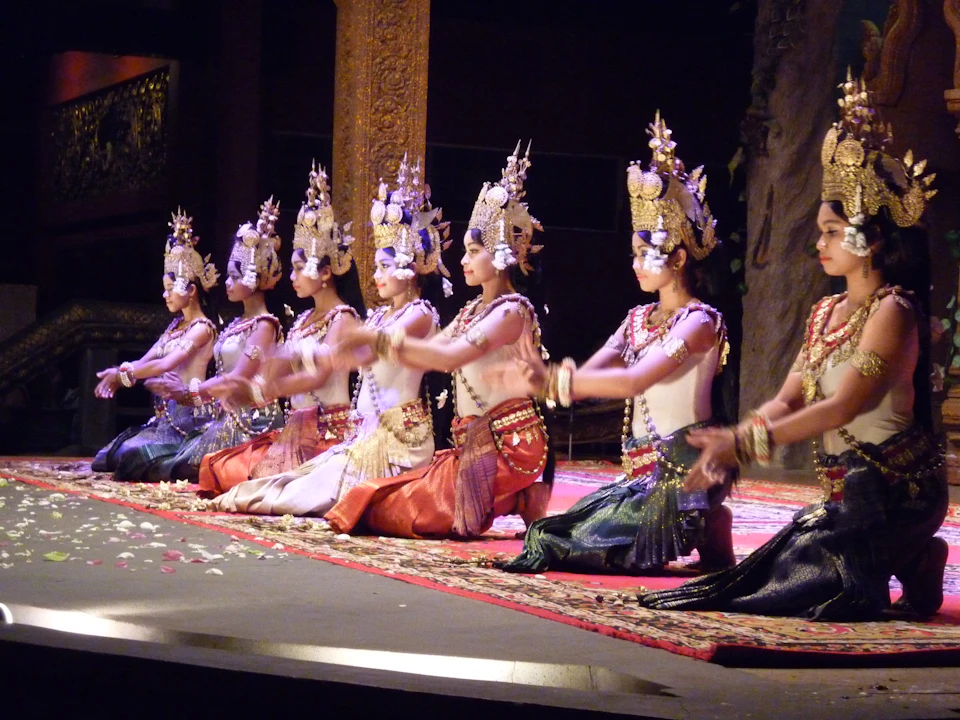 Several women, seated in a line on stage, wear traditional ornate headdresses and costumes, performing a synchronized dance.