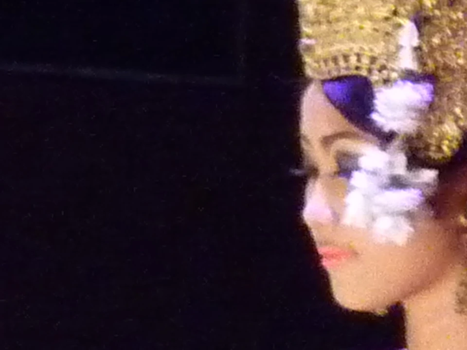 Several women, seated in a line on stage, wear traditional ornate headdresses and costumes, performing a synchronized dance.