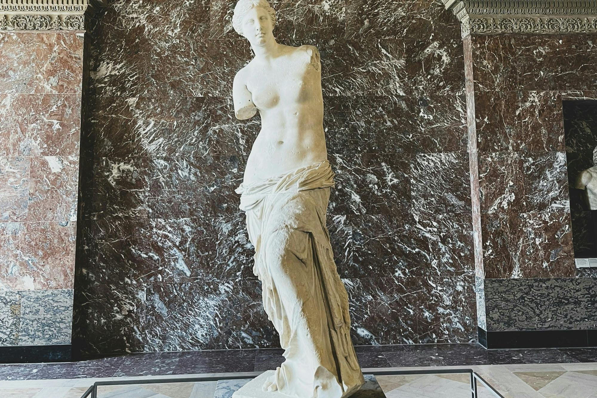A marble statue of a classical figure without arms, draped in fabric, displayed against a dark marble wall.