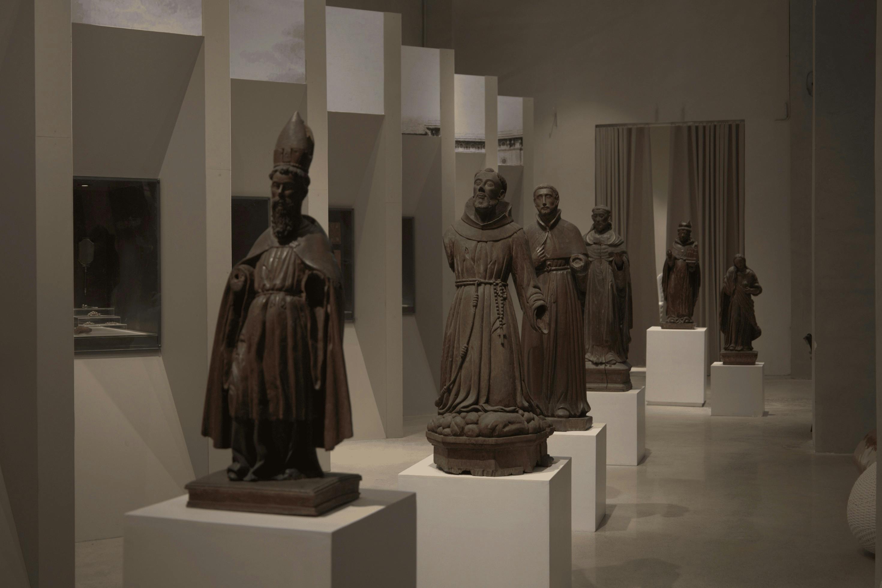A museum exhibit featuring statues of religious figures on pedestals in a modern, minimalist setting.