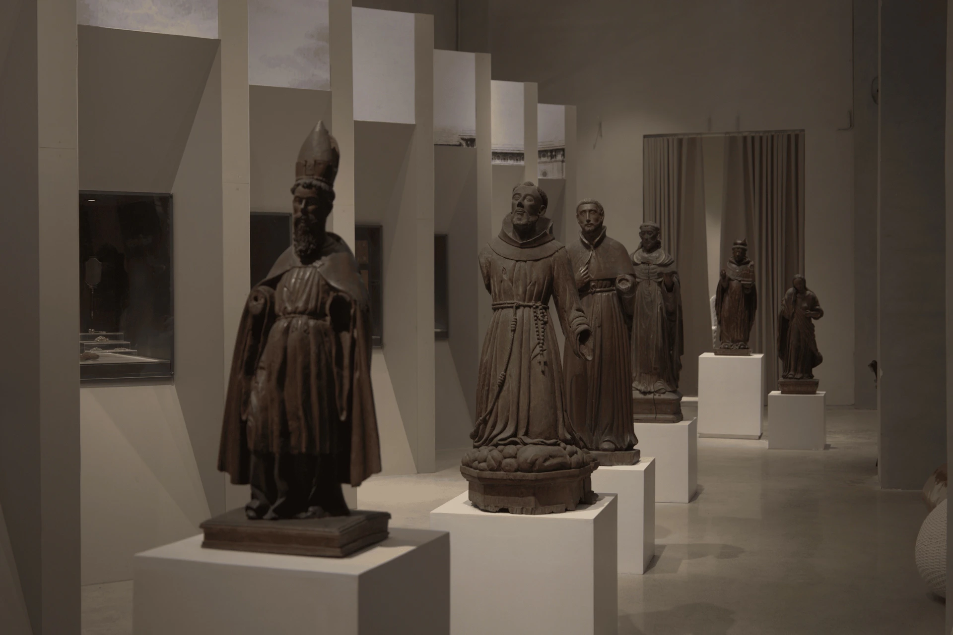 A gallery displaying wooden religious statues on pedestals under dim lighting, arranged in a row.