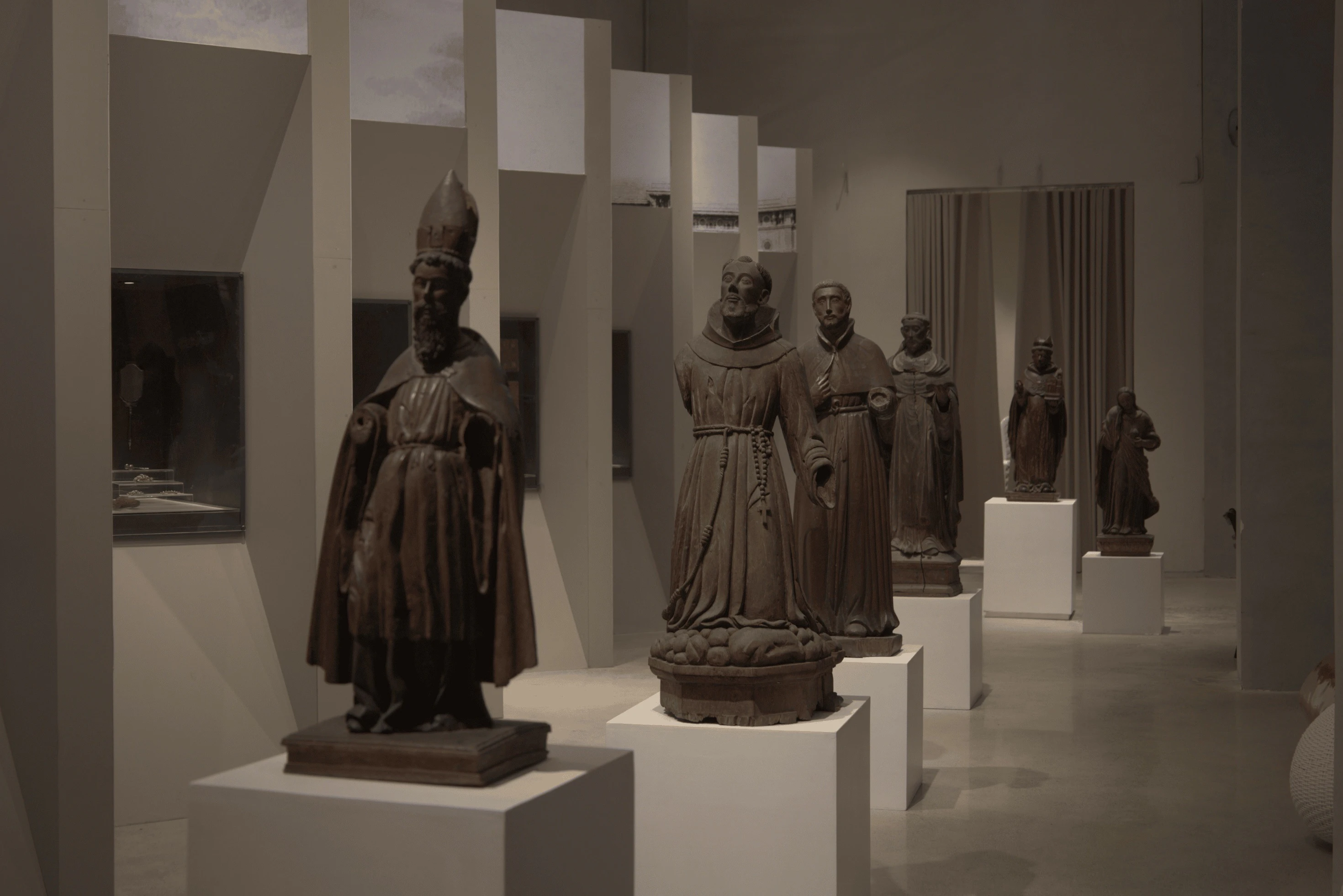 A gallery displaying wooden religious statues on pedestals under dim lighting, arranged in a row.