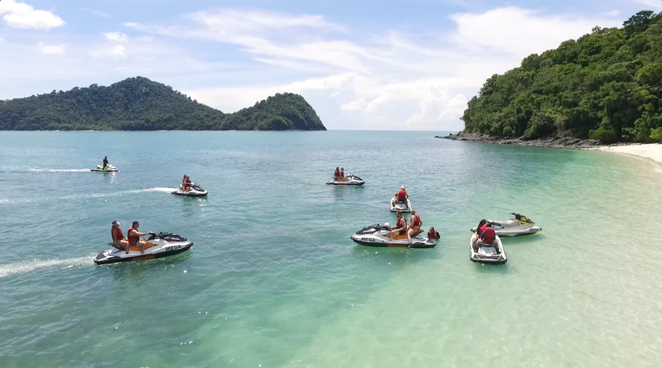 People riding jet skis on a clear turquoise sea, surrounded by green hills under a partly cloudy sky.