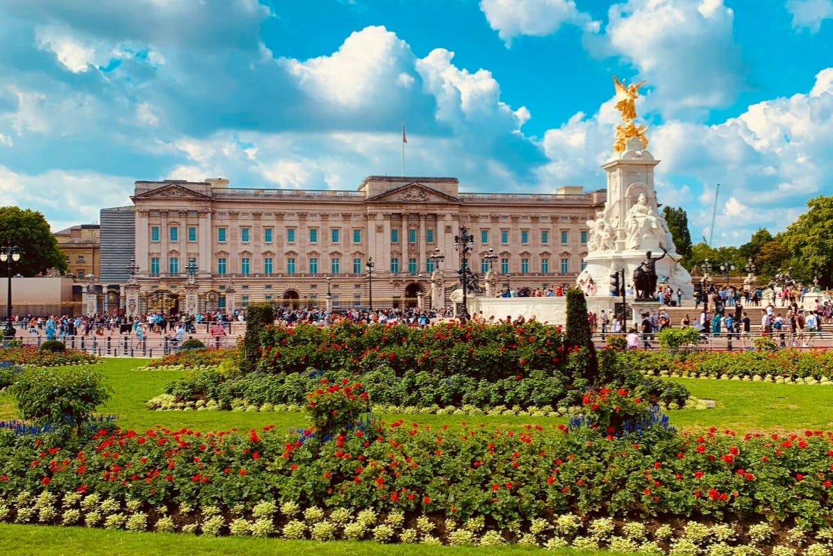 A grand building with a flag, a statue and fountain in front, and colorful flower gardens in the foreground under a blue sky.