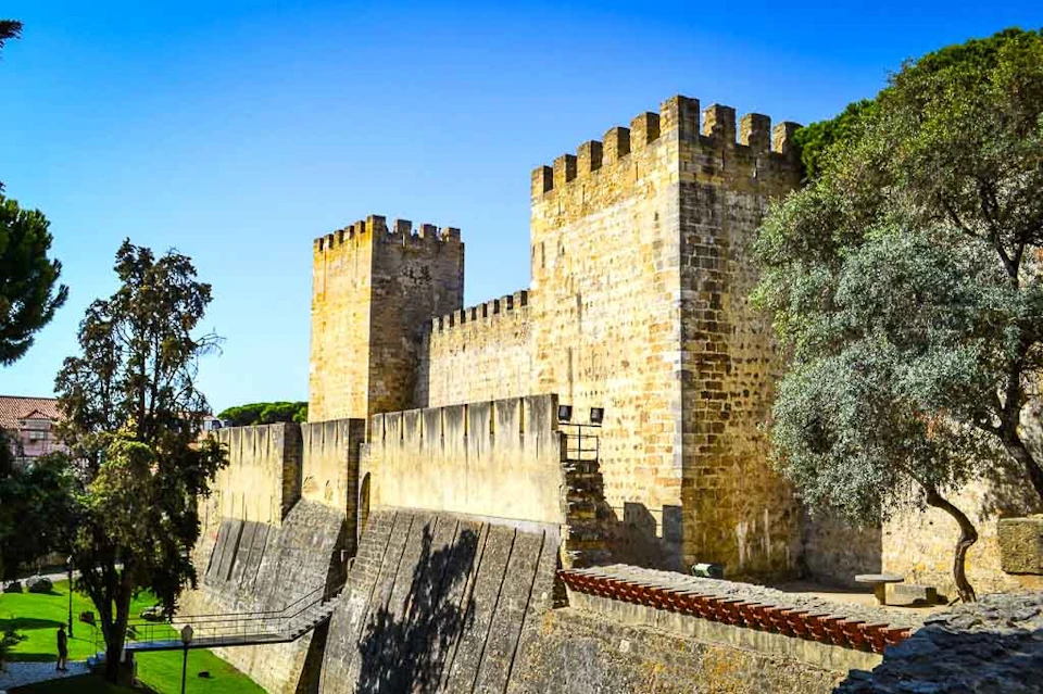 Ancient stone fortress with two square towers and crenellated walls surrounded by trees under a clear blue sky.