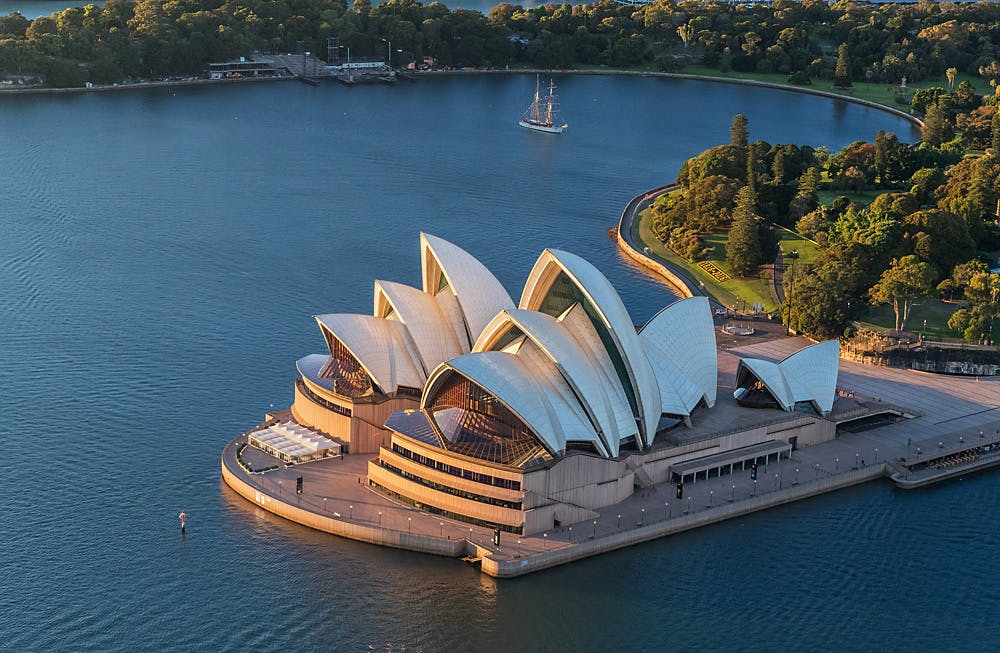 Vue aérienne de l'Opéra de Sydney avec ses voiles blanches emblématiques, entouré d'eau et de verdure, avec un voilier à proximité.